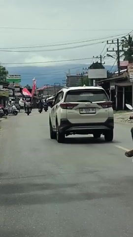 A white SUV is driving down a street. A person on a scooter is driving behind the SUV. A white truck is driving behind the scooter. The street has buildings and trees on either side. The sky is cloudy.