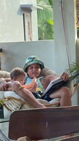 A man wearing a camouflage bucket hat sits in a hanging chair with a baby in his lap. The man is reading a book to the baby and pointing at the pictures. The baby is looking at the book. The chair is on a porch with a concrete wall and a plant in the background. The man has a tattoo on his arm.