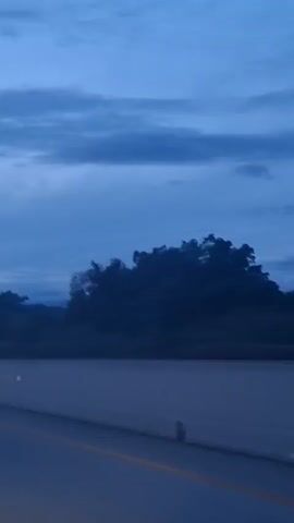 The video displays a vertical shot from a moving vehicle, looking out at a landscape under a cloudy, dark blue sky. The foreground features a road or barrier with a yellow line marking its edge. In the mid-ground, silhouetted trees and foliage are visible against the sky. The background consists of dark, hazy mountains. The overall impression is of a twilight or early morning scene during possibly overcast weather. There are no visible people or distinct objects in the frame besides the natural landscape and the road.
