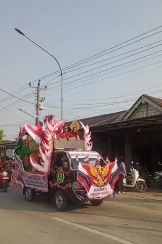 A black pickup truck decorated for a parade is driving down a street. The truck is adorned with red and white streamers, flags, and an emblem of the Garuda Pancasila. On the truck bed, there are people in traditional Indonesian attire and a large, colorful, dragon-like decoration. Two people are seated in the passenger seats of the truck cab. The parade appears to be a patriotic celebration.