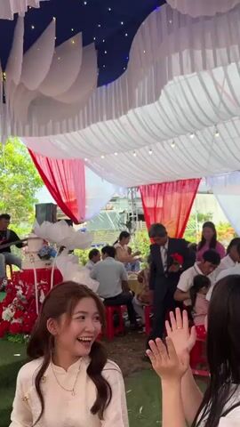 The video shows a wedding celebration under a tent. 
There is a woman in a white traditional dress, smiling and clapping. A man in a suit walks by with a red flower. Two musicians play instruments to the left of the screen. A woman in a red dress sings. A large screen in the background shows photos of the wedding couple. The tent is decorated with white and blue draping, white flower decorations, and strings of lights. Several guests are present.
