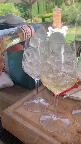 The video begins with a close-up shot of a person's hand pouring a clear liquid from a bottle labeled with red lettering into two wine glasses filled with ice. The glasses are positioned on a wooden board outdoors, with a blurred background of a patio or garden. As the liquid is poured, it mixes with the ice. The camera then pans slightly to the right, revealing a third wine glass, also filled with ice, and a blurry background that includes a white umbrella, green trees, and a brick structure. The lighting suggests it is daytime.