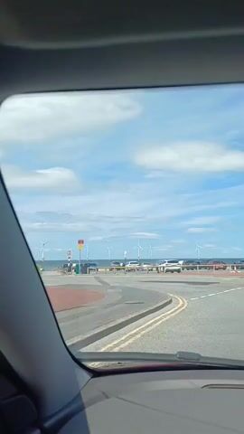 The video shows a view from inside a car driving on a road. The road curves to the left. In the distance, there is a body of water, likely the sea, and a line of wind turbines. Several cars are parked or driving along the road beside the water. The sky is blue with white, fluffy clouds, suggesting it is daytime and the weather is fair. The sun appears to be shining. The scene gives the impression of a coastal drive, possibly in Redcar, UK, as suggested by the wind turbines which are a prominent feature of the Redcar coastline. There is no visible text in the video. The overall mood is relaxed and summery, depicting a casual journey on a pleasant day.