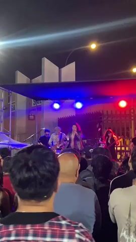 A live music performance takes place outdoors at night. The stage is set up on a metal scaffolding structure covered by a blue awning, with lighting arranged in red and blue. Onstage, a lead vocalist sings into a microphone while backed by musicians playing instruments, including a guitar. The audience stands in front of the stage, holding up phones to record the performance. The crowd is diverse in appearance. In the background, buildings are visible under streetlights.