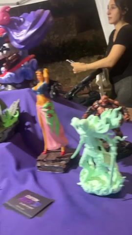 A panning shot reveals a table covered in a purple cloth displaying various character figurines. The figurines appear to be from popular anime and comic series, including Dragon Ball Z, and possibly Marvel and Star Wars. To the right of the frame, a woman with dark hair and wearing a black top is partially visible, appearing to interact with the display. The background is dimly lit, suggesting an outdoor or indoor event at night. Several figurines are prominent, including a large green and black figure with red hair and large circular hands, a purple figure with a cape, a blonde female figure in a bikini-like outfit with a pink and green wrap, and a muscular green-haired figure. Smaller figurines and accessories, such as miniature cars and busts, are also present. Some of the figurines have price tags displayed in front of them. The overall impression is of a merchandise display at a convention or a similar event.