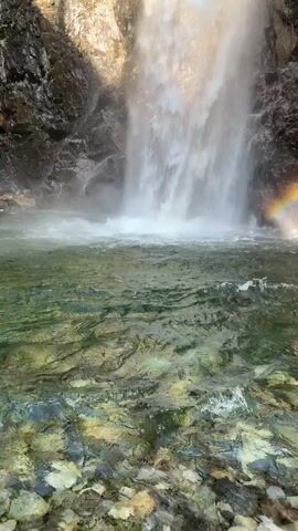 This is a close-up, upward-angled video of a waterfall cascading down a rocky cliff face. The water is misty and turbulent at the bottom, with sunlight creating a rainbow effect in the spray. The water plunges into a clear, shallow pool with a rocky bottom. Lush green trees and vegetation are visible at the top of the cliff. The lighting suggests it is daytime. There are no people or animals visible in the video.