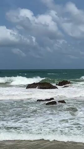 The video shows a choppy ocean with waves breaking against rocks near the shore. The sky is partly cloudy. The camera is positioned at a low angle, looking out towards the sea.