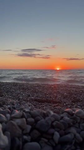 The video is a low-angle shot of a rocky beach at sunset. The sun is setting over the ocean, casting an orange glow across the sky and water. The waves are gently lapping at the shore, and the pebbles in the foreground are wet and glistening. The sky is a clear blue with a few wispy clouds. The overall mood is peaceful and serene.