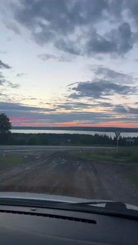 The video shows a view from inside a vehicle, looking out at a rural landscape during what appears to be dusk or dawn. The sky is filled with clouds, some of which are illuminated by soft pink and orange light near the horizon, suggesting the sun is low. A body of water, possibly a lake, stretches across the background, bordered by a dense line of trees. In the foreground, a dirt road or intersection is visible, with some puddles indicating recent rain. A triangular yield sign stands on the right side of the road. The interior of the car, including the dashboard, is partially visible at the bottom of the frame. The camera is steady, and the overall impression is of a serene, natural setting.