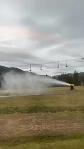 A snow cannon sprays out a plume of artificial snow onto a grassy field. Overhead is a ski lift, its chairs unoccupied. In the background are green mountains under a cloudy sky.
