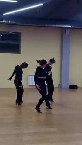 Four women are in a dance studio, rehearsing a choreography routine. They are all wearing black outfits. The studio has light yellow walls, a wooden floor, and a window. The dance includes movements like hip sways, shoulder rolls, and arm gestures.
