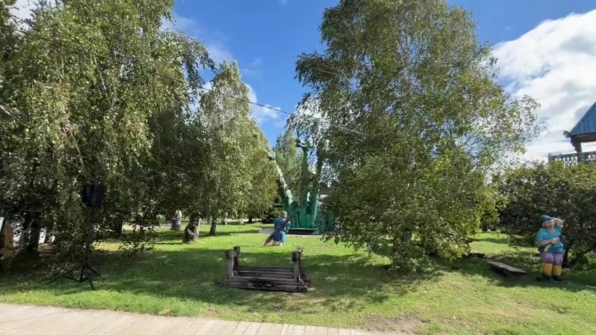 The video begins with a wide shot of a park-like area with trees and grass. A black speaker on a stand is visible on the left. In the distance, a large green dinosaur sculpture is present. Further on, a wooden structure resembling a gazebo or tower stands. The video pans to the right, revealing a path and more trees. A wooden structure with a peaked roof and balconies is prominently featured. Beyond it, a wide river and lush green landscape are visible under a partly cloudy blue sky. The camera then moves to show a rustic wooden building with stairs leading to its entrance. Green fences and decorative wooden figures are also present. The video ends with a shot of the wooden building and its surroundings. No people are clearly visible in the video.
