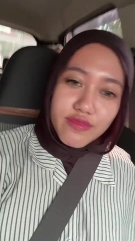 This media file features a young woman wearing a dark maroon hijab and a white and black striped collared shirt. She is seated in a car, with a grey seatbelt visible across her chest. The background shows the interior of the car and a blurred view of the outside, suggesting movement.

The woman's facial expressions change throughout the video. She begins with a neutral expression, then smiles broadly, revealing braces on her teeth. Finally, she makes a pouty face. Her makeup includes pink lipstick and visible eyeliner.

The activity appears to be a short recording or selfie video taken by the woman while in transit. The lighting suggests it is daytime, possibly late morning or afternoon, and the outdoor environment is not detailed enough to determine weather conditions. The setting is a car interior, and the presence of a seatbelt indicates the vehicle is likely on a road. There is no visible text in the media file.