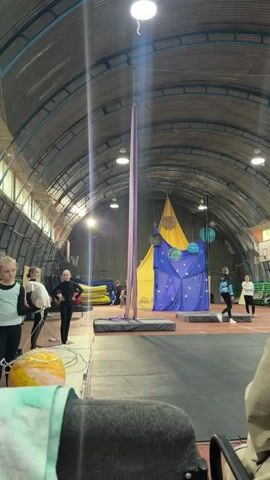 The video displays an indoor gymnasium or performance space, likely for aerial arts. A tall, purple aerial silk hangs from the ceiling, anchored by a rigging system. In the background, a vibrant stage setup is visible, featuring a yellow draped canopy behind a blue fabric backdrop adorned with white stars and several hanging spherical decorations resembling planets. Two padded mats are placed on the floor in front of the aerial silk, likely for safety.

Several individuals are present in the scene. On the left, two young women are standing near the aerial silk. One, in a black outfit, appears to be a performer, possibly demonstrating a pose. Another, in a white and black top, stands nearby. Further to the right, two more women are conversing near the stage. In the background, other figures can be seen, suggesting a group activity or practice session.

The lighting suggests it is daytime, with artificial lights illuminating the space. The overall atmosphere seems to be one of preparation or practice for a performance or training session. The setting is utilitarian, with exposed metal beams and a corrugated metal roof, typical of a gymnasium or an older sports facility. No visible text is present in the frame.