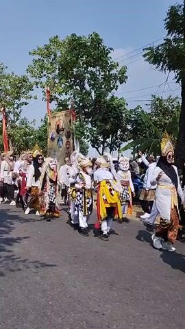 A group of people dressed in traditional clothing and costumes are participating in a parade on a paved road under a clear blue sky. Trees line the right side of the road, and a banner with pictures is visible in the background. Some individuals are wearing elaborate masks and headdresses, while others are in simpler traditional attire. The people appear to be walking or dancing in a procession.
