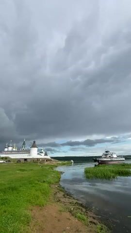 A wide shot shows a body of water in the foreground, with a sandy bank and green grass on the left. A white building complex with green domes, likely a monastery, is visible on the left bank. A ferry is moving on the water on the right side of the frame. The sky above is filled with dramatic, dark storm clouds. There are a few cars parked near the monastery. No people are clearly visible. The overall atmosphere is overcast and potentially stormy.