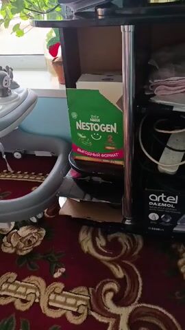 The video pans up and to the left to show a child's walker on a red rug patterned with roses. The walker is white and gray, with a gray seat and a tray holding colorful toys. The camera moves around the walker, revealing a green box of Nestogen 2 baby formula sitting on a shelf next to the walker. There is also a window in the background with a plant on the windowsill.