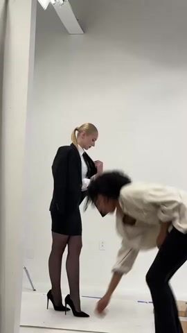 A woman in a black suit jacket, white shirt, black skirt, black stockings, and black high heels stands with her hands near her jacket. To her right and slightly in front, another person with curly dark hair is out of focus, crouching down. The scene is in a room with white walls and a white floor.