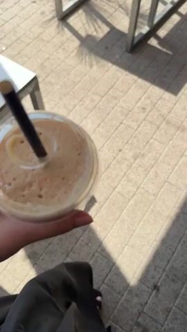 The video begins with a close-up of a person's hand holding a clear plastic cup filled with a frothy beverage and a black straw, placed outdoors on a tiled surface. The camera then pans to reveal an outdoor seating area with white-topped tables and metal legs, some with ashtrays. Palm trees and potted plants with red flowers are visible, along with a glimpse of a road, grass, and distant mountains under a clear blue sky. The overall environment appears to be a cafe or restaurant with an outdoor seating section.