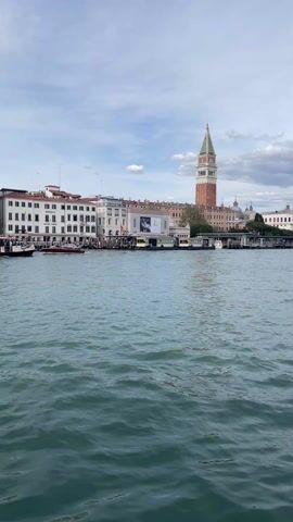 The video depicts a waterway scene in Venice, Italy, taken from a low angle. The water is choppy, with small waves. Along the waterfront, there's a line of buildings, predominantly white with red rooftops. Piers extend into the water, and several boats are docked. In the distance, the tall St. Mark's Campanile stands prominently against the sky. The sky is partially cloudy, with patches of blue visible.
