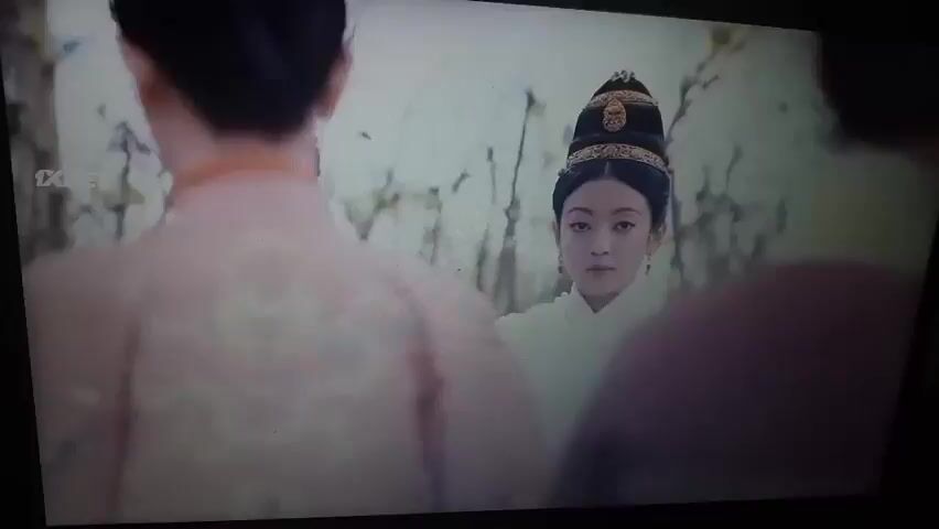 The video shows a scene from what appears to be a historical drama, likely set in ancient China, based on the clothing and hairstyles. In the foreground, a young woman with elaborate hair and traditional attire is visible. She is looking directly at the camera. Behind her, other figures in similar historical clothing are blurred and out of focus, suggesting they are in the background or moving. The setting seems to be outdoors, possibly in a garden or courtyard, with greenery and natural elements suggested. A logo that says "1XBET" is visible in the upper left corner of the screen. The scene transitions through several frames, showing slight shifts in the camera angle and the positions of the people. In some frames, the woman is looking down or to the side, and in others, she is looking towards the viewer. The overall impression is of a staged historical scene with a focus on the central female character.
