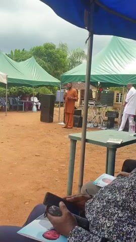 Here's a description of the video:

The video shows an outdoor event taking place in a sandy, open area, possibly in a village or rural setting. Two men are standing near a table set up with sound equipment, including speakers. One man is dressed in an orange outfit and is speaking into a microphone. The other man, in a white suit, is holding a book or papers. Several tents are set up in the background, under which people are sitting on chairs.

The video is shot from the perspective of someone sitting under a blue-and-white umbrella. A person wearing a floral shirt and holding a phone is visible in the foreground. A book and a bottle of water are on the table beside them. The weather appears overcast.