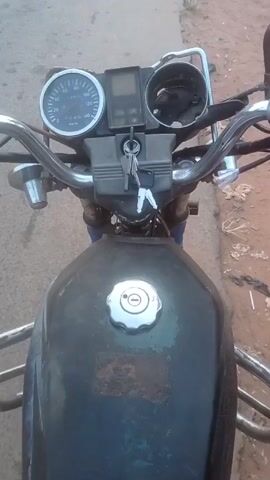 The video shows a point-of-view shot from the seat of a motorcycle. The motorcycle is stationary on a paved road. The dashboard is visible, showing a speedometer and a digital display, with keys in the ignition. The gas tank is in the foreground, with a silver cap. To the right of the road is a dirt shoulder. In the distance, a person is visible near a corrugated metal structure. The weather appears to be sunny.