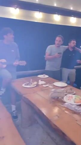 A group of men are gathered outdoors under a covered area with string lights overhead, around a rustic wooden table laden with food. On the table are plates with food remnants, bread, plastic cutlery, and bags of salad. In the foreground, a man on the left is playing an acoustic guitar. To his right, a man in a grey t-shirt holds a phone and gestures upwards, seemingly in mid-conversation or action. Another man in a black t-shirt stands behind him, also gesturing. Further to the right, a man in a black hoodie with a white brace on his arm is holding a drink and looking towards the center. The scene appears to be a casual outdoor gathering with food and drinks.