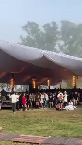 The video shows a crowd of people at an outdoor music festival. There's a large white tent-like structure with stage lighting and speakers inside. People are dancing and walking around. The ground is covered in grass and some wooden planks. In the background, there are trees and other structures, including white tents and a decorative golden arch. The atmosphere is foggy and the sky is overcast. A couple is sitting on the wooden planks, watching the scene.