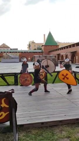 This media file shows a historical reenactment or live-action role-playing event taking place outdoors on a wooden platform. The subjects are several people dressed in what appears to be medieval or Viking-inspired attire, including helmets and armor. Two individuals in the foreground are actively engaged in a mock sword fight, each wielding a sword and a shield. One fighter is in a low, lunging stance, while the other stands more upright.

In the background, two other individuals are present, also holding shields. One appears to be standing still, while the other might be observing or preparing to join the fight. The setting is an open courtyard, possibly within a historical fort or museum complex, given the presence of brick buildings with arched windows and a prominent tower with a green, conical roof in the distance. Wooden barricades and some stacked wooden materials are also visible. The ground is covered with grass.

The time of day appears to be late afternoon or early evening, with soft, diffused light, suggesting it's either dusk or an overcast day. The weather seems calm, with no strong winds apparent.

There is some visible text on a banner in the far background, but it is mostly illegible due to distance and resolution. The overall atmosphere suggests a controlled, recreational event focused on historical combat. The participants appear focused and engaged in their activity.