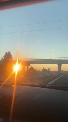 A first-person perspective video shows a car driving on a highway at sunset. The sun is visible as a bright orb on the left side of the screen, casting lens flares. A bridge passes overhead. The sky transitions from a clear blue to orange and yellow hues near the horizon. Trees and distant signs are visible along the roadside. The interior of the car, including the dashboard and windshield wipers, is partially visible in the foreground.