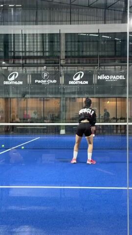 A person in athletic wear is playing padel on a blue court. They are holding a padel racket and are in the motion of hitting a ball. The court is enclosed by glass walls and metal fencing. In the background, other people are visible on adjacent courts, and there are various advertisements and signs around the facility. The lighting appears to be artificial.