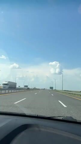 A first-person perspective video shows a car driving on a highway under a clear blue sky with scattered white clouds. The highway has multiple lanes marked with white lines. Streetlights are visible on both sides of the road. In the distance, other vehicles can be seen. The car is moving forward, and the view from the windshield shows the road stretching ahead. Green signage is visible on the right side of the highway. The sun is shining, creating a bright environment. The dashboard of the car is partially visible in the foreground.