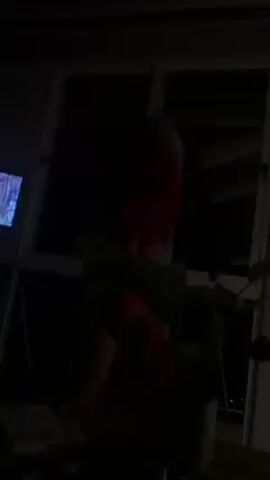 The video shows a person standing just outside a sliding glass door. The person is partially obscured and is wearing a red shirt and light shorts. They are holding a green towel over their head. The outdoor area is dark. High-pitched, electronic-sounding music plays.
