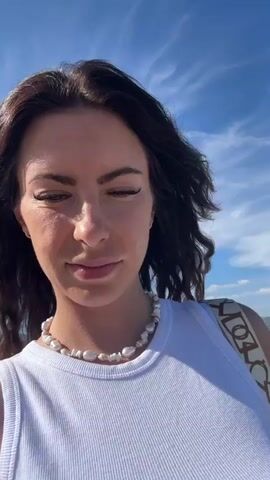 The video shows a woman standing on a beach, looking at the camera. She has long, dark, wavy hair and is wearing a white tank top and a pearl necklace. Her facial expression is neutral with a slight smile. In the background, the beach is visible with sand, a few people, and the sea. Further in the distance, there are buildings and hills, suggesting an urban or resort area. The sky is blue with some white clouds. The lighting suggests it is daytime, likely late morning or afternoon, with bright sunlight. A bag with a geometric pattern is visible on her right shoulder. A tattoo of a rose is visible on her left arm. The overall scene conveys a relaxed, summery atmosphere. There is no visible text in the video.