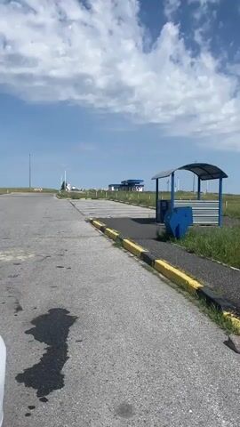 The video shows a view of an open, grassy field under a cloudy blue sky. A paved road or pathway is visible in the foreground, with yellow and black markings along its edge. To the right of the pathway, there is a small, covered structure with blue siding. Beyond the immediate foreground, the field is dotted with evenly spaced posts, and further in the distance, there are more posts and what appear to be wind turbines or similar structures. A building with a blue roof and a white tower is visible in the far distance to the left.