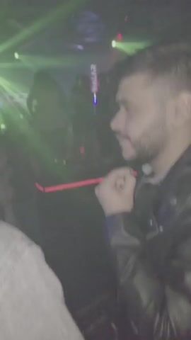 The video shows the interior of a nightclub. Green spotlights shine across a dance floor where several people are standing, some dancing. At the back, a screen displays the word "YALAR" vertically in red. A DJ booth is visible with a computer screen displaying audio waveforms. A man in a gray denim jacket is seen drinking from a bottle of beer. The lighting is dim, and the atmosphere appears lively.