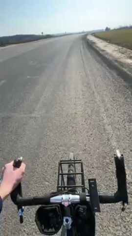 The video shows a first-person perspective of someone riding a bicycle on a rural road. The road is gray and appears to be made of gravel or a similar material. The sky is blue and clear. On either side of the road there are fields of what appears to be farmland. The bicycle is equipped with a rack on the back, and pouches on the front. The person is wearing a jacket with blue and red stripes on the sleeve. The video is slightly shaky, likely due to the movement of the bicycle.