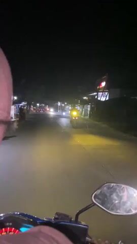The video shows a first-person perspective of someone riding a motorcycle at night on a street. The rider is wearing a pink headscarf and a pink shirt. The street is paved and lined with trees and buildings, some of which have lights on. There are other motorcycles and vehicles on the road, with their headlights illuminating the scene. The overall atmosphere is dark and appears to be a residential or commercial area at night. The video quality is somewhat blurry due to the movement and low light conditions.