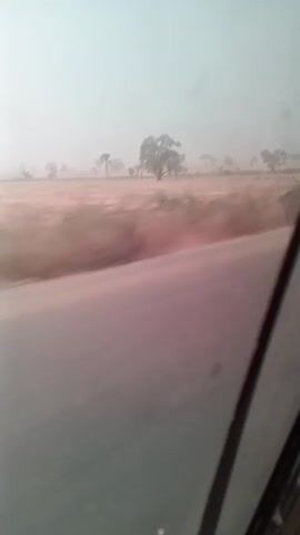 The video shows a view from a moving vehicle looking out the side window at a rural landscape. The landscape is flat and dry, with sparse vegetation and a few scattered trees visible in the distance. The sky is hazy, and the overall visibility is low.  The road adjacent to the landscape is gray and appears to be paved. The vehicle's window frame is visible on the right side of the frame, slightly obscuring the view.