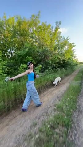 A young woman with long brown hair is walking down a dirt path in a rural area, with a white dog following behind her. The woman is wearing a blue tank top, baggy jeans, a cap, and flip-flops. She is holding a bunch of flowers or weeds in her hand and is twirling around as she walks. The dog is walking calmly behind her. The path is surrounded by green trees and grass on either side. The sky is blue and clear.