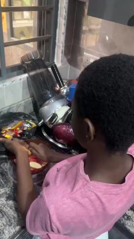 A person with dark skin and short, dark hair, wearing a pink t-shirt and white shorts, is standing in a kitchen. They are holding a knife in their right hand and a piece of red fruit, likely an apple, in their left hand, and appear to be cutting or peeling it. The fruit is in a clear plastic container on a speckled countertop. To the left of the person, there is a black trash bag with some items inside. Behind the fruit and trash bag, there is a blender and a metal pot or bowl. A window with metal bars is visible in the background. The overall environment suggests a domestic setting, likely a kitchen counter, with evidence of food preparation.