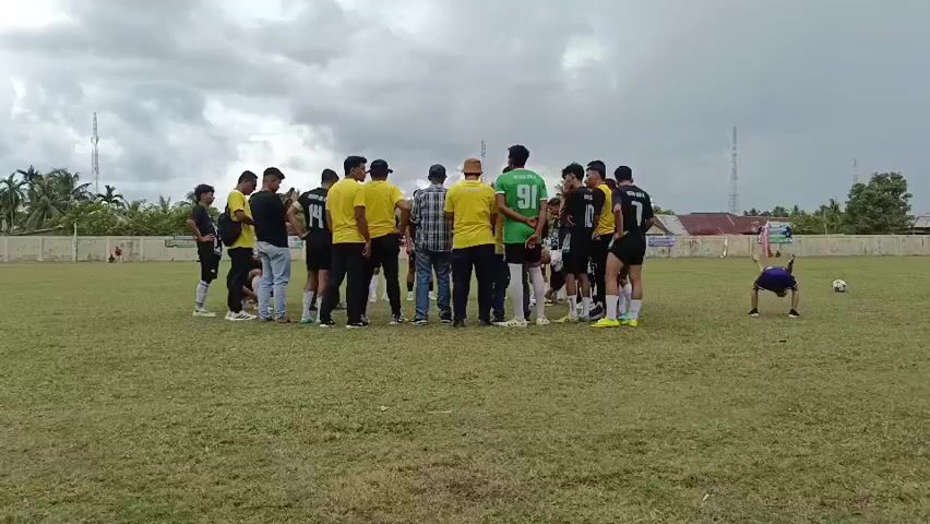 A group of about fifteen young men, mostly wearing yellow shirts and black shorts, are gathered in a circle on a grassy field, seemingly engaged in a discussion or pep talk. Some of the men are wearing jerseys with numbers. A few adults are also present in the circle, one wearing a green shirt and another in a plaid shirt. In the background, a fence runs along the edge of the field, and beyond that, there are buildings and trees. To the right of the circle, a younger boy in a blue shirt is visible running with a soccer ball. The sky is overcast.