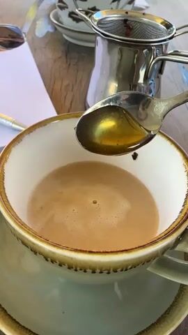 A close-up shot shows a spoon of honey being drizzled into a cup of coffee. The coffee is light brown with foam on top. The spoon is metallic and reflects the light. The background is blurred, but it appears to be an outdoor setting with a wooden table and a tea strainer.