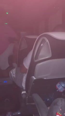 The video shows the interior of a bus, possibly a party bus, with people riding in the back. A man wearing a pink t-shirt and white shorts is standing between the rows of seats. Another person is sitting in a seat, wearing a white shirt and gray Nike shorts. A sound system with a speaker is on the floor between the seats, and the bus is filled with red light or haze. A water bottle is visible on the floor. The man in pink shifts his weight and adjusts his stance. The camera angle is low.