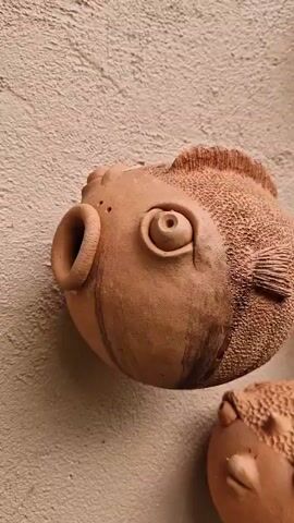 A close-up shot pans across a collection of small, earthenware fish sculptures attached to a textured wall. The sculptures are unglazed and vary in size, with detailed features such as eyes, fins, and mouths. Some fish have bulbous bodies, resembling pufferfish, while others have more elongated forms. The wall appears to be made of stucco or concrete, with a rough, uneven surface. The lighting is soft, casting gentle shadows and highlighting the texture of both the sculptures and the wall. The overall impression is one of a decorative display of handmade art.