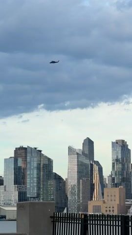 The video shows a cityscape with modern skyscrapers. A helicopter is visible flying in the sky above the buildings. The sky is cloudy and appears to be overcast. The buildings are made of glass and metal, reflecting the light. The camera angle is from a lower vantage point, looking up at the skyline. The overall impression is one of an urban environment under a dramatic sky.