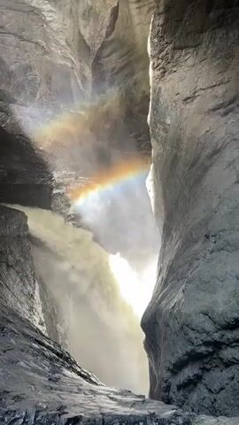 This is a video of a powerful waterfall cascading through a narrow canyon. The sheer, grey rock walls of the canyon frame the scene, with sunlight streaming in from above. The intense spray from the waterfall creates a misty atmosphere, and a vibrant rainbow arcs across the mist, illuminated by the sunlight. The water itself is a churning white mass as it plunges downwards, creating a dramatic and awe-inspiring spectacle. The textures of the wet rock and the frothing water are palpable. The overall mood is one of natural power and beauty, with a touch of magic from the presence of the rainbow. The time of day appears to be daytime, with the sun positioned high enough to illuminate the canyon. No people or man-made objects are visible, emphasizing the raw, untamed nature of the location. The location context provided indicates this is in Interlaken, Switzerland, a region known for its dramatic alpine scenery and waterfalls. There is no visible text in the video.