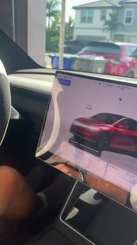 A person's hands are visible on the steering wheel of a car. The steering wheel has a white top and a textured dark gray bottom. The car's dashboard features a large touchscreen display showing a navigation map and vehicle information. The screen is illuminated and angled towards the viewer. Outside the car, a residential street is visible with houses, palm trees, and other vehicles parked. The lighting suggests it is daytime, possibly sunny. The video appears to be shot from the driver's perspective, looking at the steering wheel and the dashboard. The person is wearing shorts and a bracelet on their left wrist. A blue trash bin is visible in the background.
