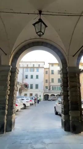 The video shows a view through an arched entryway into a courtyard. The courtyard has a paved ground and is surrounded by buildings. Several cars are parked in the courtyard. A few people are visible, one person walking in the center of the courtyard, and two people standing near a parked car on the left side. The sky above is blue with some wispy clouds. The architecture suggests a European setting.